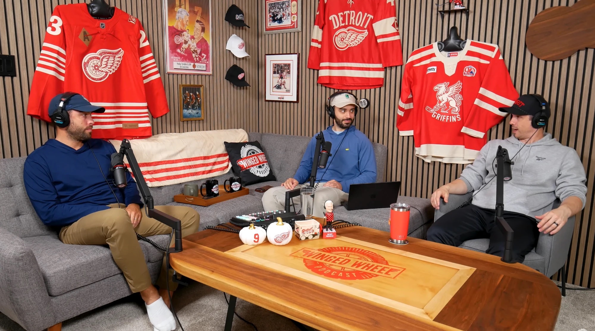 Ryan, Evan and Brad in the WWP studio, seated on the couch with microphones and headphones, Red Wings jerseys and framed photos on the wall behind them